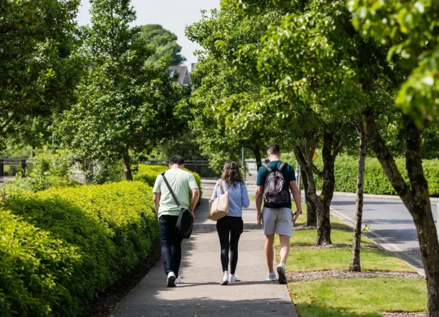Three students, two boys and a girl, walking away from the camera through the ATU Sligo Campus in the sunshine and surrounded by leafy green trees.