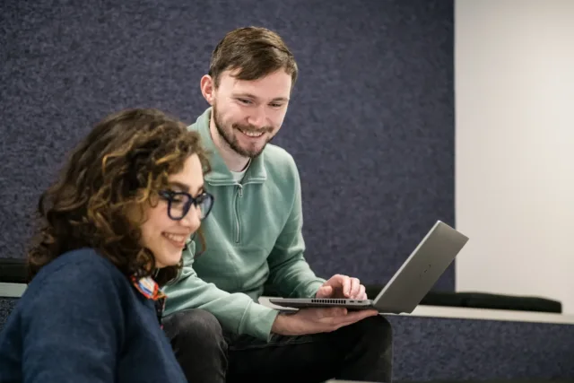 Male and female student laugh while looking at laptop screens