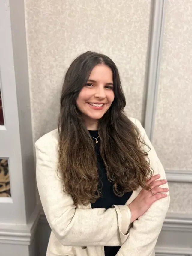 Female student in a cream top with long brown hair standing against a wall with her arms crossed