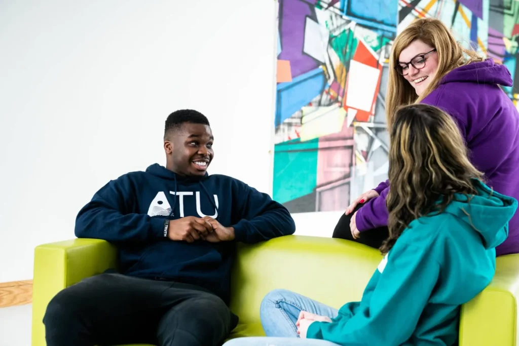 Three people sit and stand around a low seating area indoors, facing each other in conversation, with one person wearing a sweatshirt displaying the text “ATU” and a colourful abstract wall artwork in the background.
