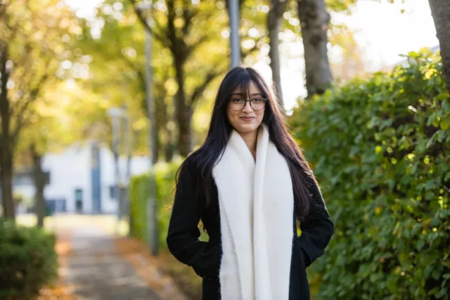 Female international student with large white scarf stands with hands in pockets along autumnal pathway.