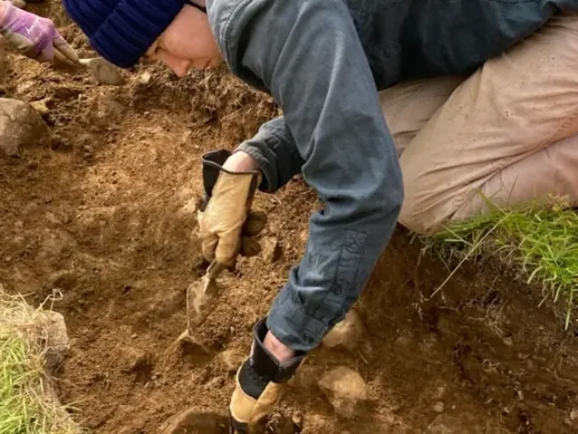 Student is knelt down digging at an archaeological excavation.