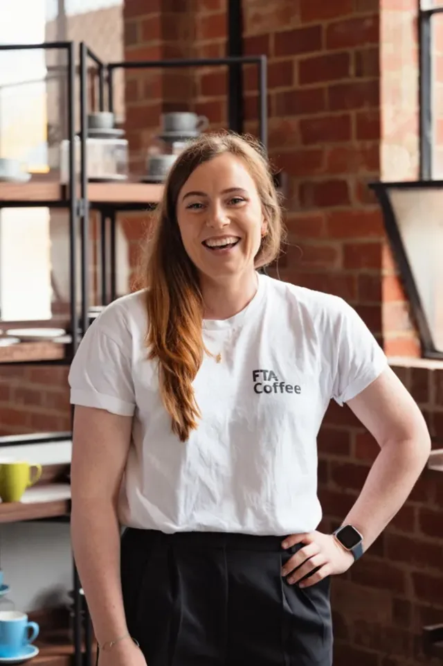 Women with red hair poses infront of red brick. She is wearing a white t-shirt with the logo FTA Coffee