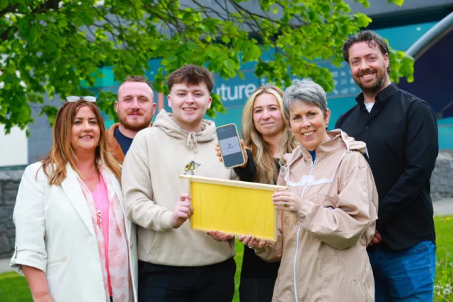 Group photo of three students, two lecturers, and the ATU President in a beekeeping suit, standing outdoors by a tree. A student showcases a phone with the HIVE app, and another helps hold a hive component with the President.