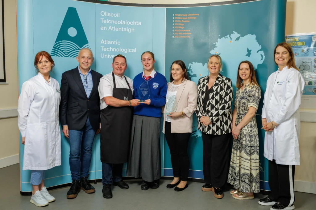 Chef Neven Maguire stands centre with the teacher on his left and the winning senior student to his left both holding glass trophies. Also in the photo is ATU staff and stakeholders, posed in front of a blue ATU branded backdrop featuring the university's logo and map of Ireland.