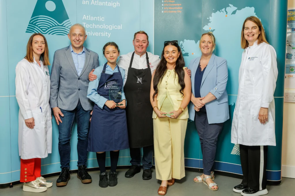 Chef Neven Maguire stands centre with the teacher on his left and the winning junior student to his left both holding glass trophies. Also in the photo is ATU staff and stakeholders, posed in front of a blue ATU branded backdrop featuring the university's logo and map of Ireland.