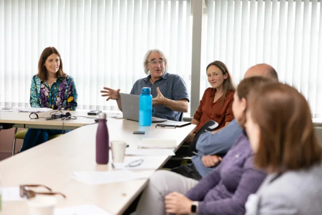 A group of people sit around an L-shaped table engaged in a discussion. One man, seated at the corner, has a laptop in front of him and is gesturing with his hand as he speaks. The others are listening attentively, focused on the conversation.