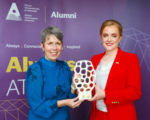 Lisa Lynch is presented an ATU Alumni Award by Dr Orla Flynn, ATU President, they both smile for the camera in front of purple alumni backdrop.