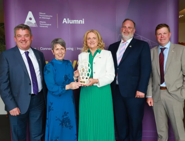 Breege Conroy Walsh is presented an ATU Alumni Award by Dr Orla Flynn and stands smiling for a group alongside ATU staff members in front of ATU alumni purple backdrop.