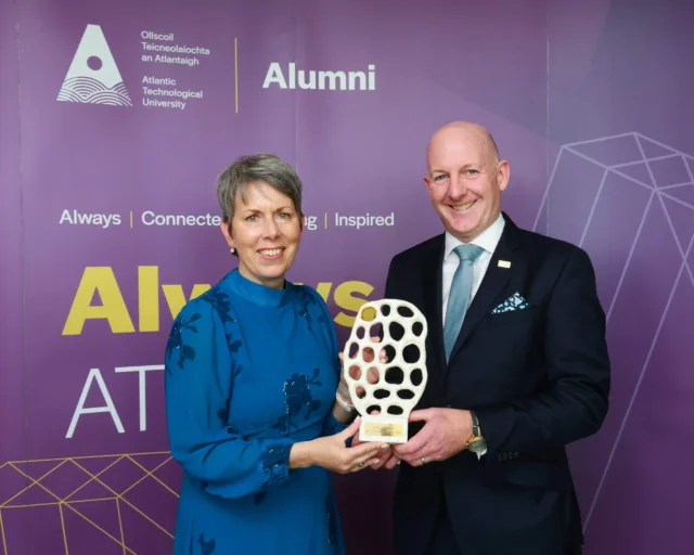 Enda McGuane is presented an ATU Alumni Award by Dr Orla Flynn, ATU President, they both smile for the camera in front of purple alumni backdrop.