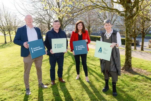 Four individuals standing on grass with trees in the background, from left to right: Dr Mark Kelly, Dr John Scahill, Dr Carina Ginty and Dr Orla Flynn. They are holding square cutouts with the following labels: 'Centre for Sustainability,' 'Education for Sustainable Development Academy,' 'Teaching & Learning Centre,' and the 'ATU Logo.