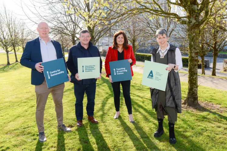 Four individuals standing on grass with trees in the background, from left to right: Dr Mark Kelly, Dr John Scahill, Dr Carina Ginty and Dr Orla Flynn. They are holding square cutouts with the following labels: 'Centre for Sustainability,' 'Education for Sustainable Development Academy,' 'Teaching & Learning Centre,' and the 'ATU Logo.
