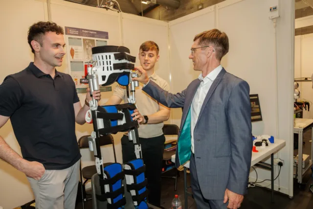 Two students, winners of the Ronan Healy Memorial Award for their project "Legatron"—a robotic device designed to assist with walking—stand alongside the Head of the Faculty of Engineering and Design. All three are examining the device.