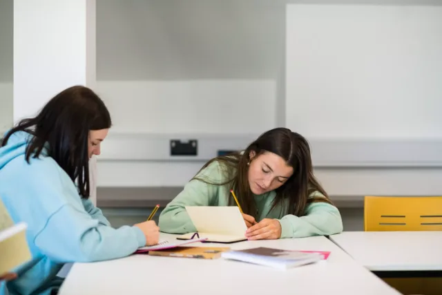 2 students are writing in notebooks whilst sat at a table