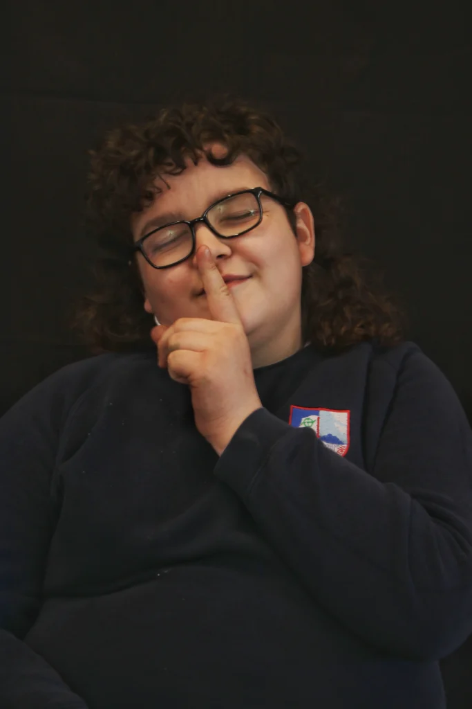 Portrait image of a pupil, wearing black framed glasses, dark hair and holds his index finger up to his mouth.