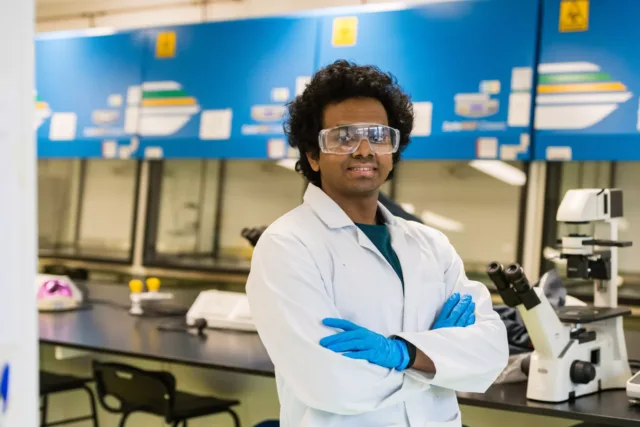 A student is stood in a lab wearing safety goggles and a white lab coat