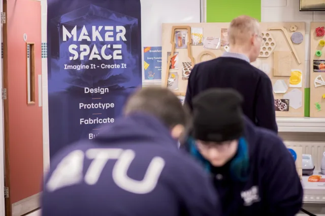 Two students with their heads down working on something. Pull up in the background that states 'Maker Space' Imagine It - Create It. There's also a man in the background looking at some projects on the wall.