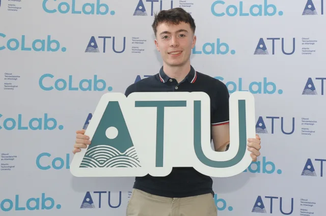 Male student holds up ATU sign smiling for camera, he is in front of backdrop with CoLab. and ATU logos across it.