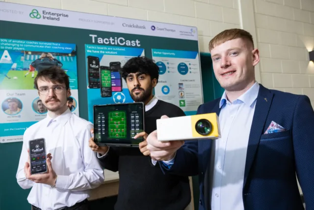 Students look at the camera holding up devices, their project TactiCast poster board is in the background
