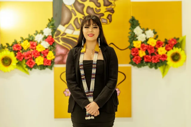 Graduate smiles for the camera wearing a black outfit and traditional jewellery and stands in front of her artist that has a yellow background and depicts a women along with flowers.