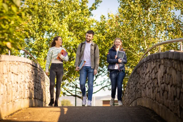 Three students walk across a stone bridge holding books and smiling engaged in conversation. The background is leafy trees.