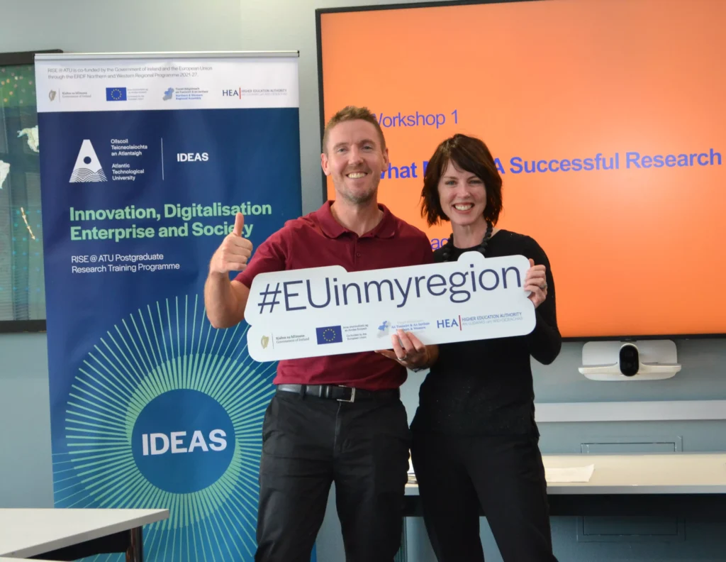 Two people smile and hold a "#EUinmyregion" sign at a research and innovation event, with an EU-themed banner and screen in the background