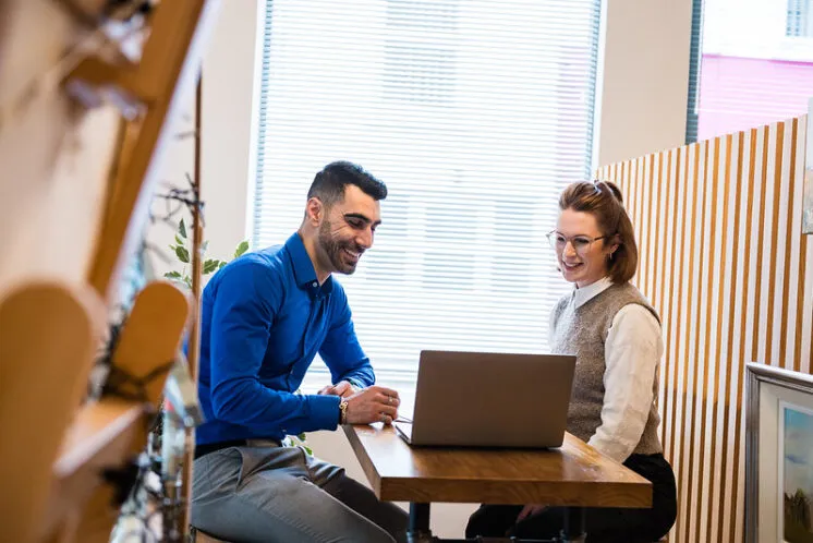 Two people sit at a small table indoors, facing each other with an open laptop between them, near a window with blinds and shelving in the background.