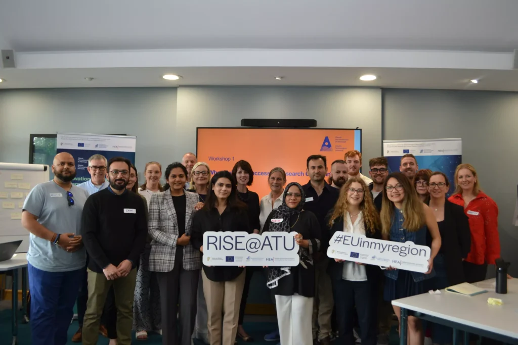 A group of people poses together at a research workshop. Two participants hold signs that read "RISE@ATU" and "#EUinmyregion,"