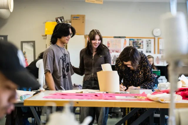 Design students working on a project. Three students are at a table covered in textiles, is marking up fabric with a pencil as the other two look on.