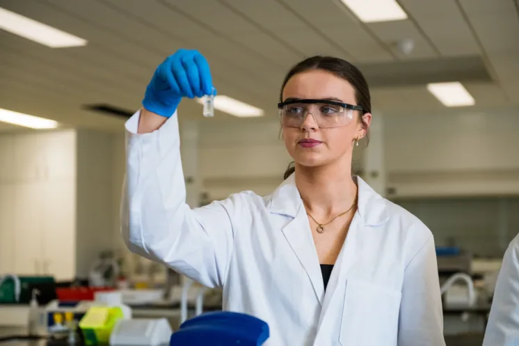 female student wearing white lab coat, safety glasses and blue gloves holds up and examines a vial in a lab setting.