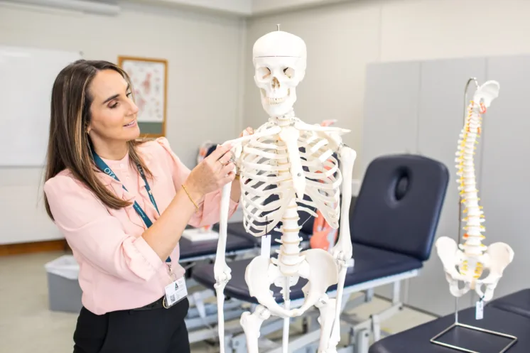 Brown haired woman in pink shirt working with a skeleton model. A model of the spine and pelvis can be seen in background.