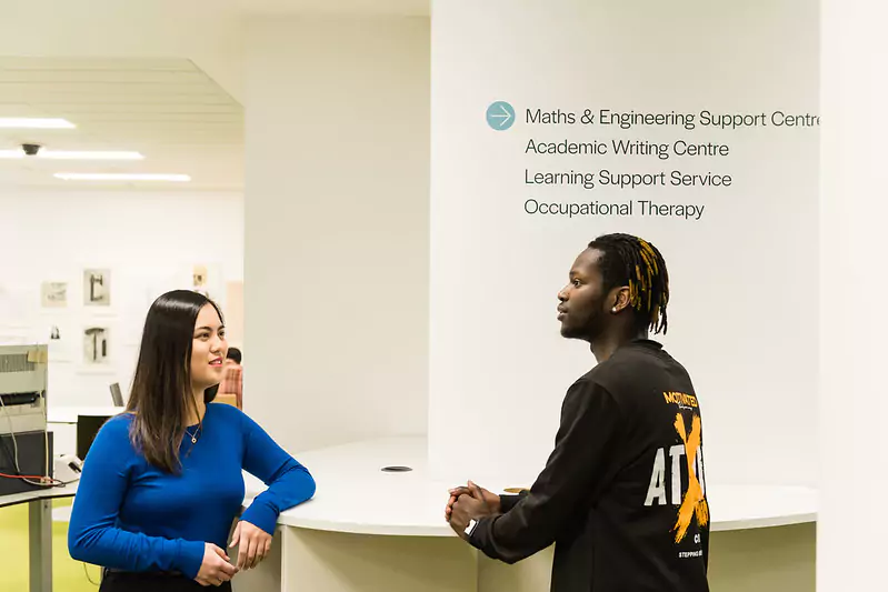 Two individuals standing and conversing near a white wall displaying text that lists academic support services: Maths & Engineering Support Centre, Academic Writing Centre, Learning Support Service, and Occupational Therapy.