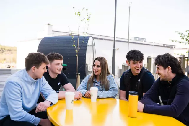 Five people sitting around a yellow table outdoors, with cups and bottles on the table. In the background, there is a modern building featuring an arched structure and small trees