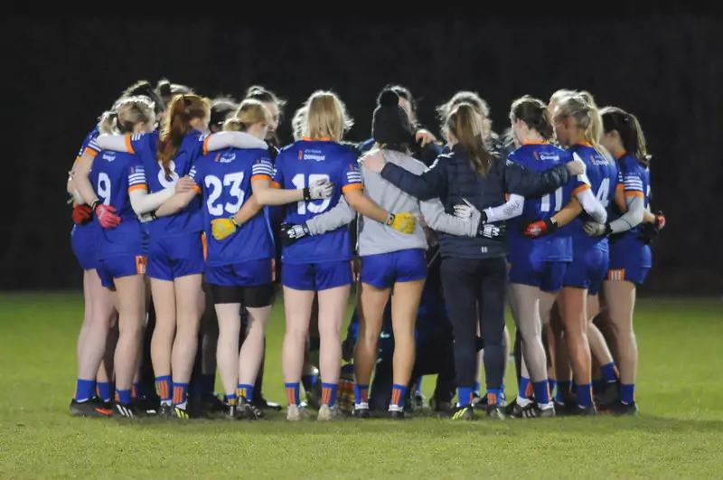 A group of people in blue sports uniforms, standing in a circle with their arms around each other on an outdoor field at night. Some individuals are wearing gloves, and uniform numbers are visible on their backs.