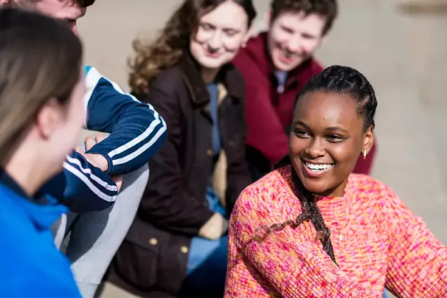 A group of people sitting together outdoors on a concrete or paved surface. They are dressed in casual clothing, including a pink and orange sweater, a blue jacket with white stripes, and various other jackets and sweaters.