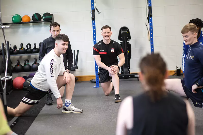 A group of people in a gym setting performing stretching exercises on the floor, including lunges. The background features shelves with kettlebells and medicine balls, indicating a fitness or training session.