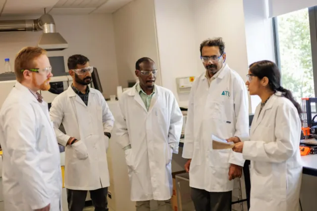 Five individuals in white lab coats engaged in discussion within a laboratory setting.