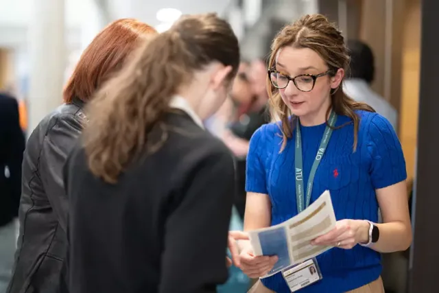 Three people standing and conversing indoors. One person in a blue shirt with a lanyard and ID badge is holding and looking at documents or pamphlets.