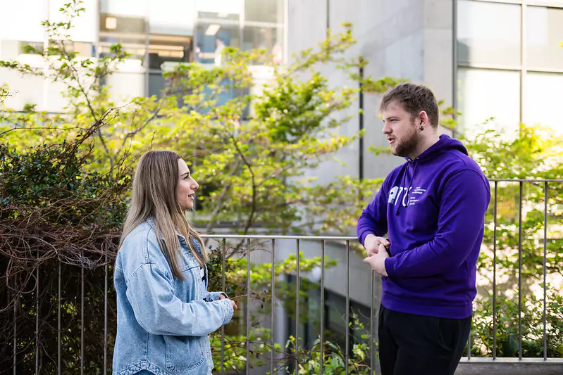 Two people standing outdoors near a railing, with greenery and a building in the background. One person is wearing a purple hoodie, and the other is wearing a light blue denim jacket.