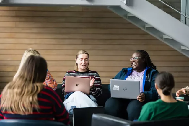 Six people sitting in a semi-circle indoors, engaged in a discussion or meeting. The setting includes wooden wall paneling and metal railings, suggesting an office or educational environment.