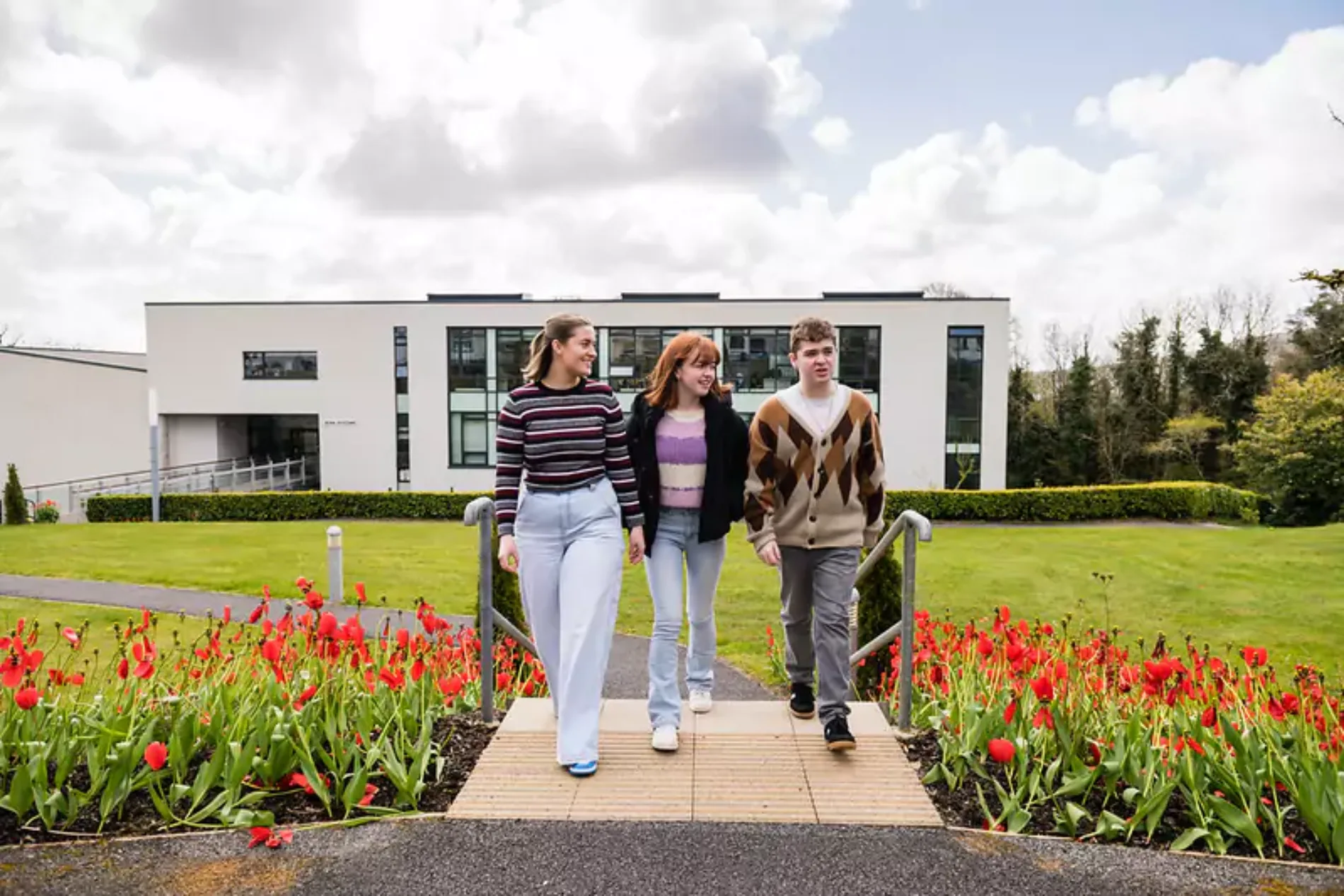 Three individuals walking along a pathway bordered by red tulips, with a modern building featuring large windows and a well-kept lawn in the background.