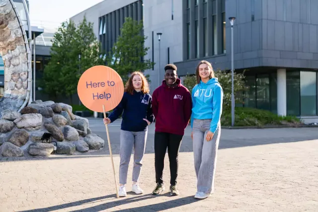 Three individuals standing outdoors in front of a modern building, wearing hoodies with 'ATU' printed on them. One person is holding a large circular sign that reads 'Here to Help'.