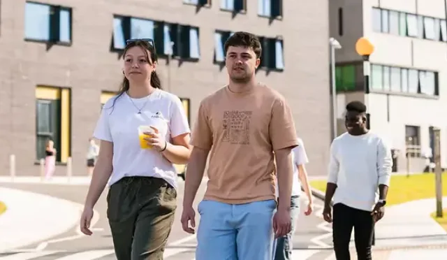 Three individuals walking on a pathway in front of modern buildings with multiple windows. One person is holding a drink, and a bicycle is parked near the building on the left. The setting includes green lawns and streetlights, suggesting an urban or campus environment.