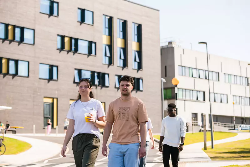 Three individuals walking on a pathway in front of modern buildings with multiple windows. One person is holding a drink, and a bicycle is parked near the building on the left. The setting includes green lawns and streetlights, suggesting an urban or campus environment.