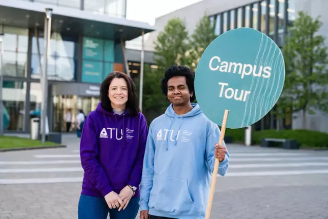 Two individuals standing outside a modern building, wearing hoodies with 'ATU' printed on them. One person in a light blue hoodie is holding a large circular sign that reads 'Campus Tour'.