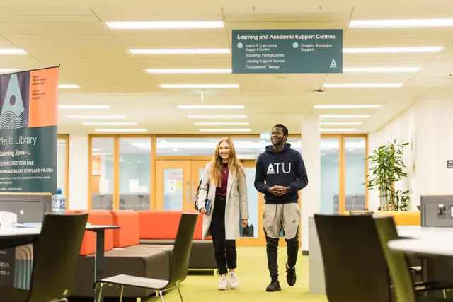 Two people walking through a library or academic support center. A sign overhead reads 'Learning and Academic Support Centres' with departments listed, including Maths & Engineering, Student Computing, Academic Writing Centre, and more. A 'Yeats Library' banner is visible on the left, and the background includes tables, chairs, and bookshelves.