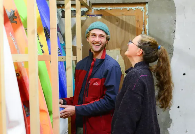 Two people standing in a storage or workshop area surrounded by colourful kayaks or similar equipment stored vertically on wooden racks.