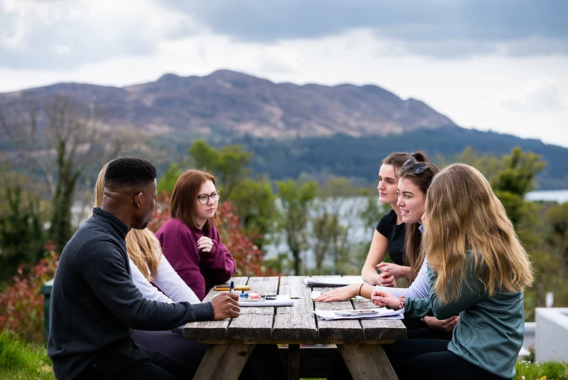 Group of students sitting on a picnic bench with notepad and pens on the table. There is a view of Lake Isle of Innisfree in background.