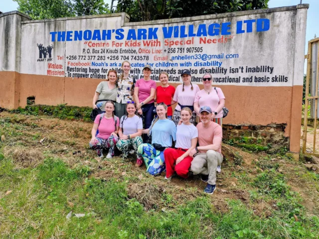 Students and Staff from ATU St Angelas visiting Uganda for humanitarian and community engagement work. Half the group is standing the other half kneels in front of them for a photo outside Noah's Ark Village which is written in blue writing on the wall behind them along with the centre's contact details, vision and mission.