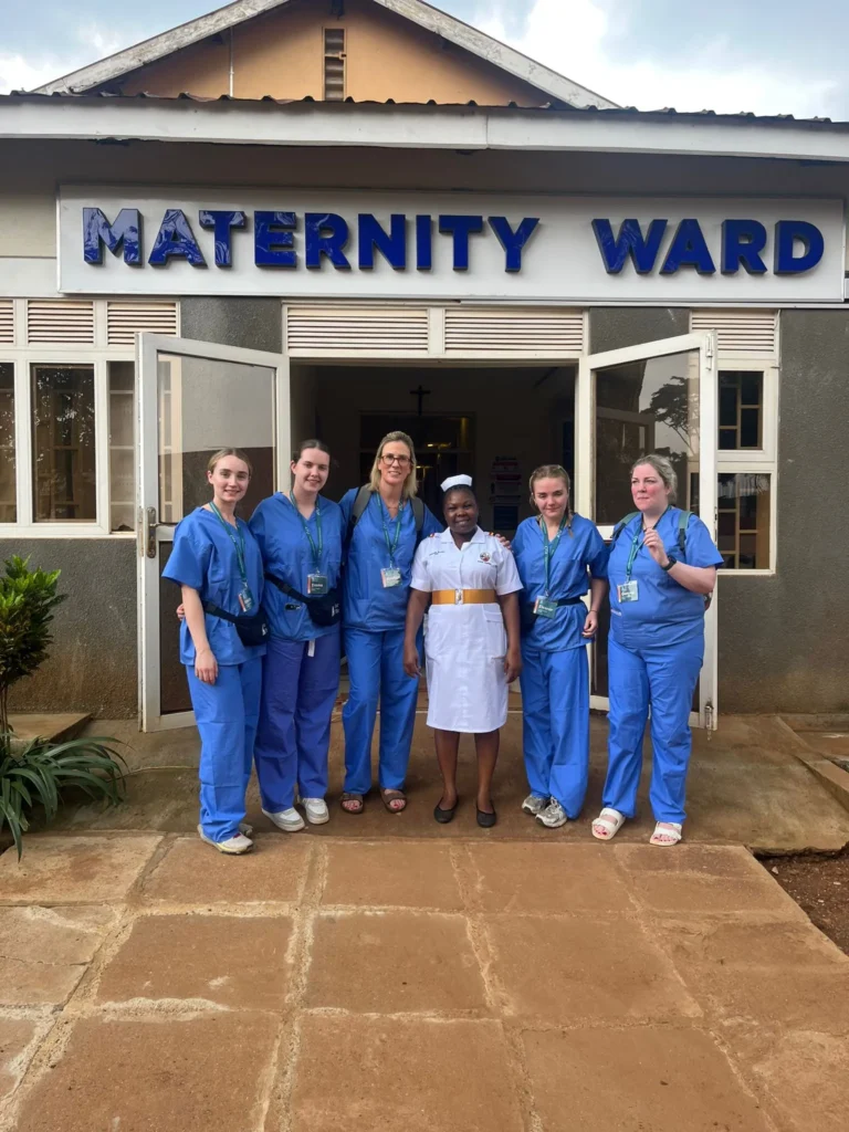ATU staff and student nurses dressed in blue scrubs stand out the front of the Maternity Ward in Uganda along with an Ugandan nurse in a white uniform in centre.
